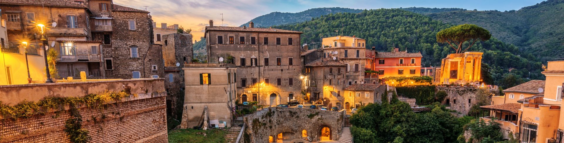 Old stone buildings sit atop a tree covered cliff side. 