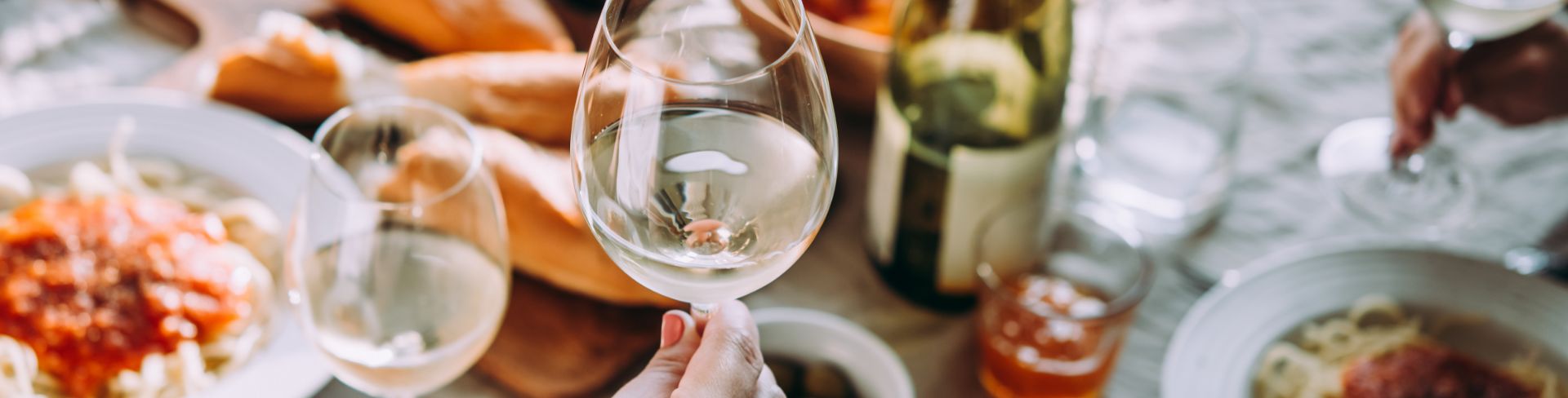 A person toasts a glass full of white wine with a table of food in the background.