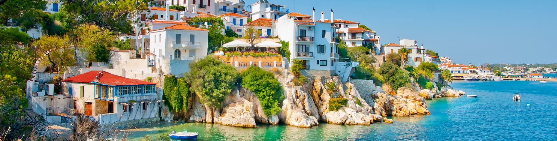 A town sits on the side of a cliff overlooking still blue waters dotted with small boats.