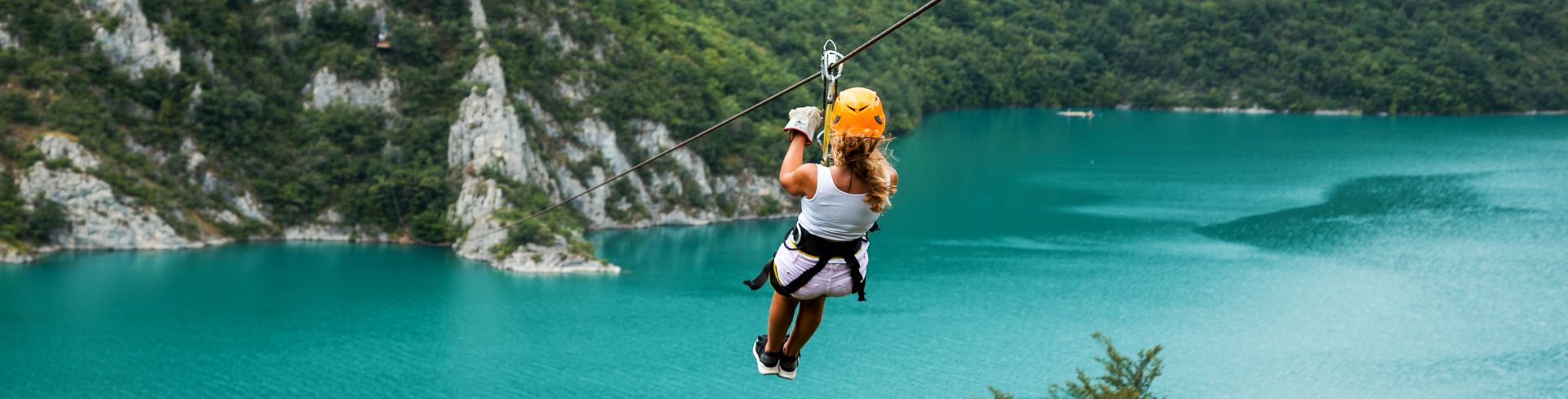 A person zip lines over deep blue waters towards a tree-covered mountainside.