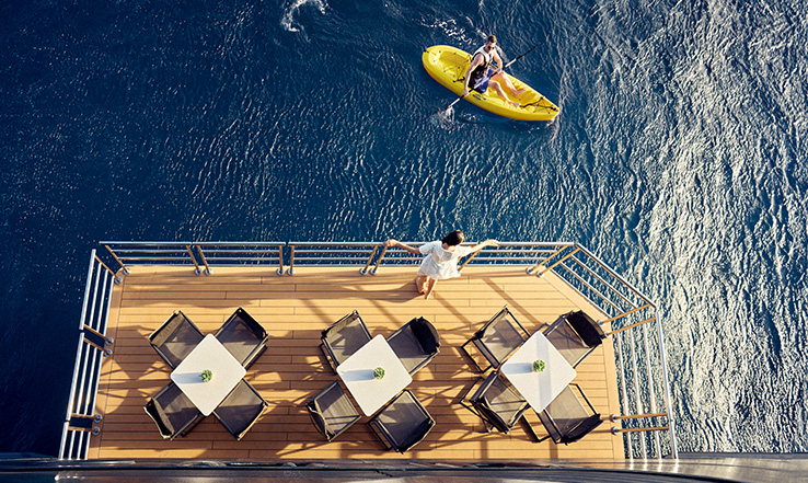 A person looks out at the ocean from a balcony while another traveler kayaks below.