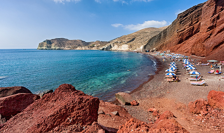 Red clay lines a beach with umbrellas and beach chairs scattered along the shoreline.