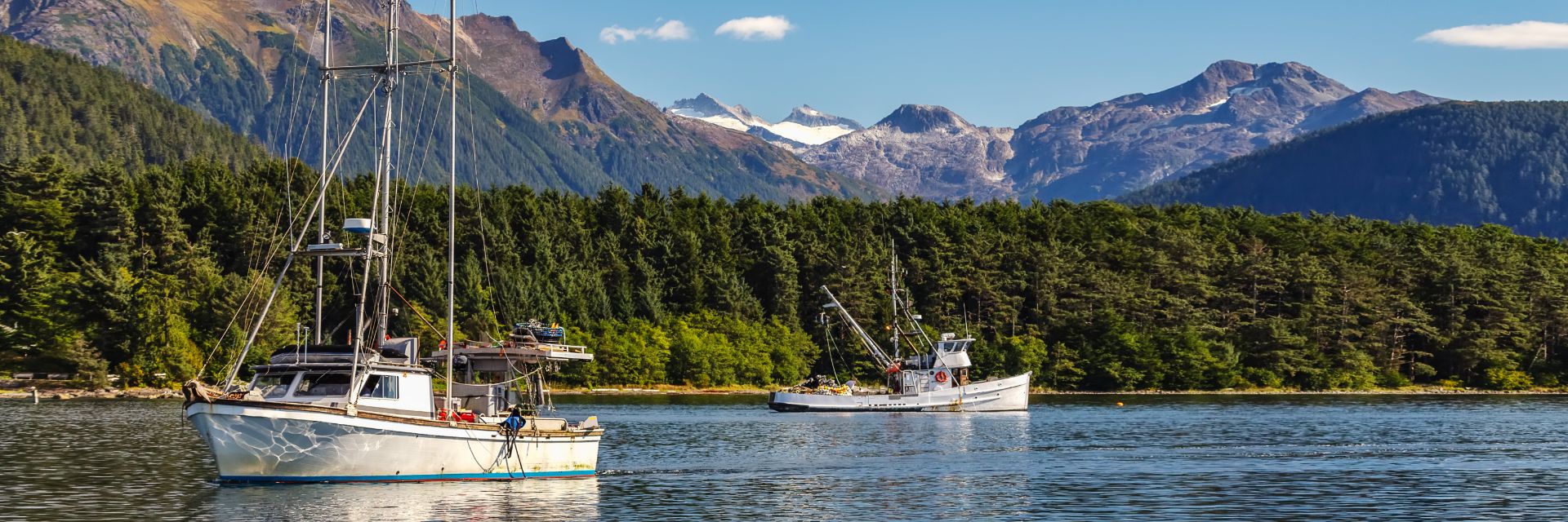 Two boats float in still waters before tree covered mountains.