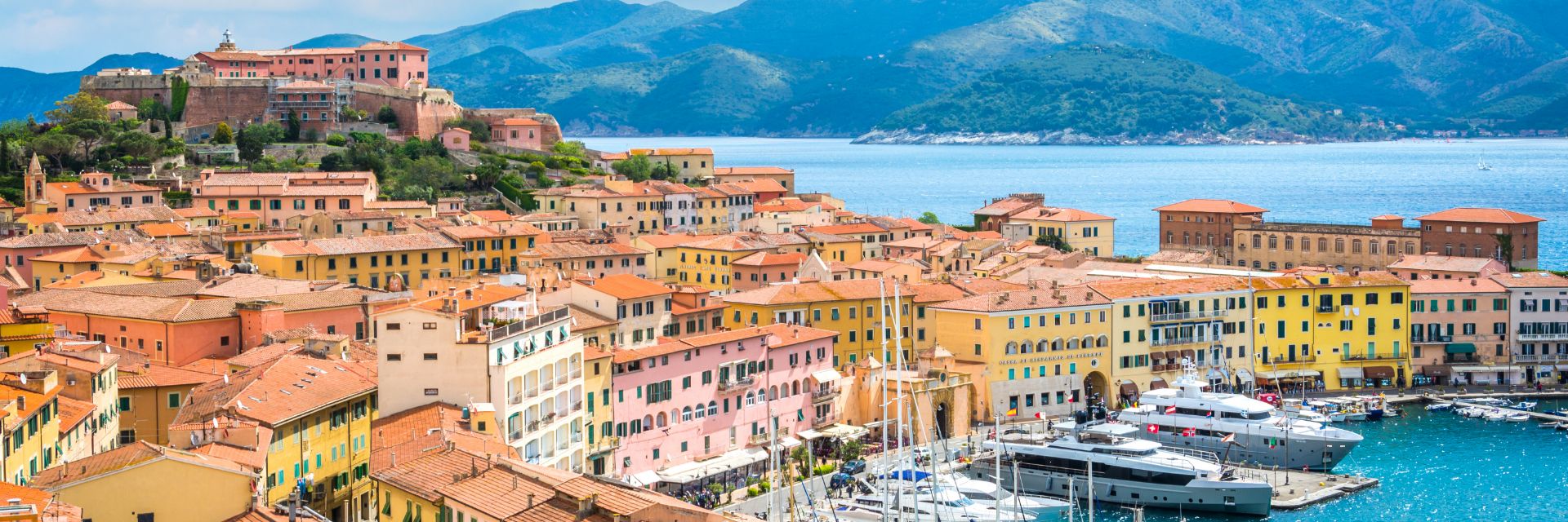 Warmly colored buildings sit oceanfront surrounded by a mountain range.