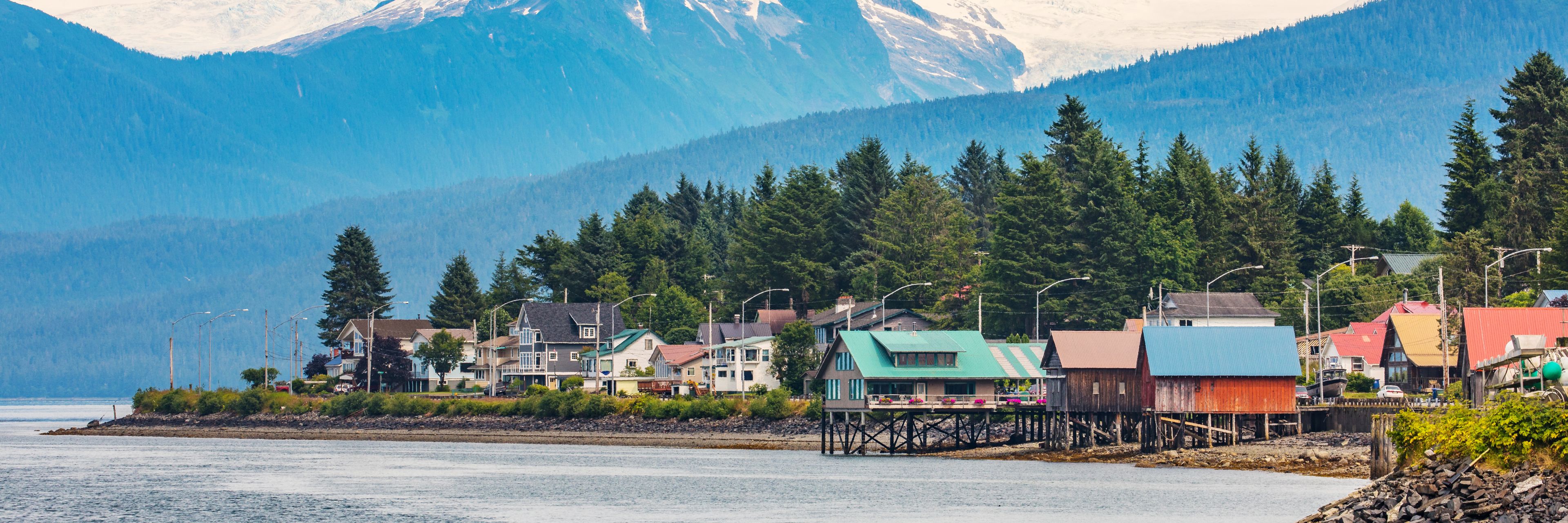 Houses line a waterway with snow capped mountains in the background.