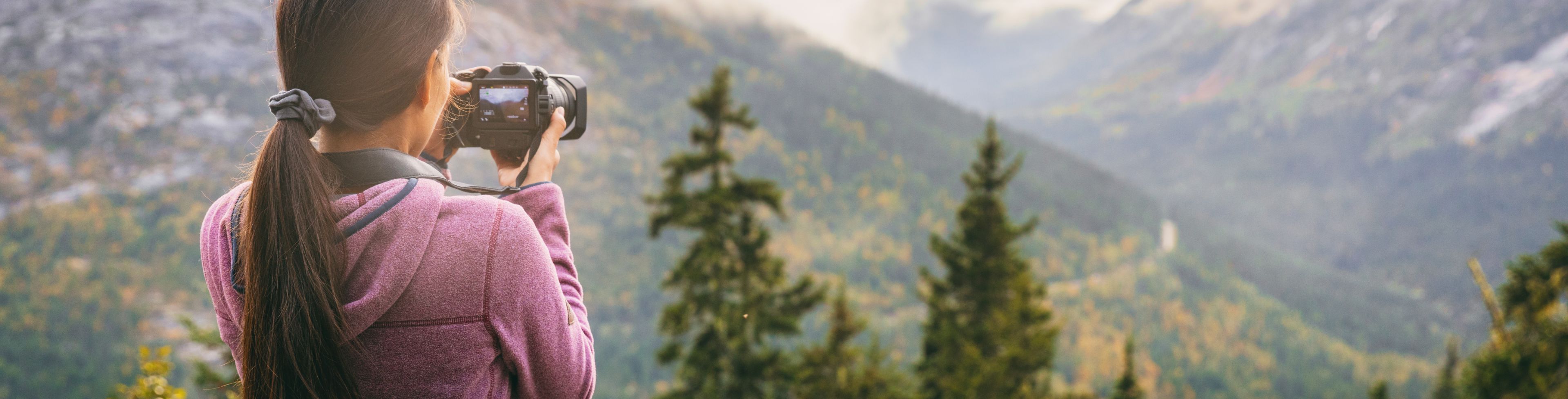 A person points a digital camera towards a tree covered mountain face.