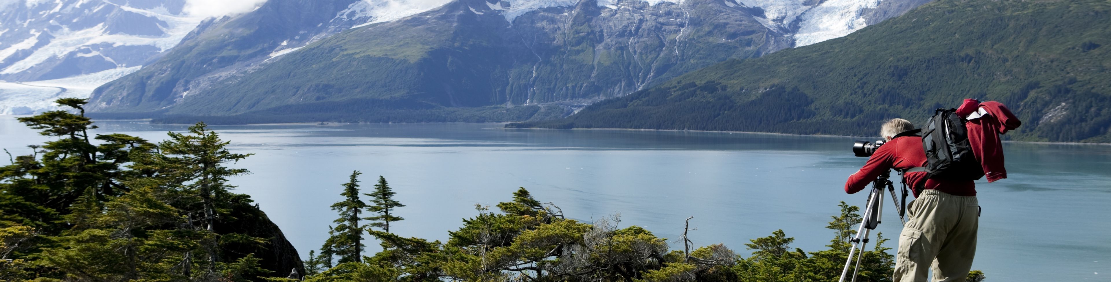 A person takes a photo of a snow capped mountain sitting behind a large waterway.