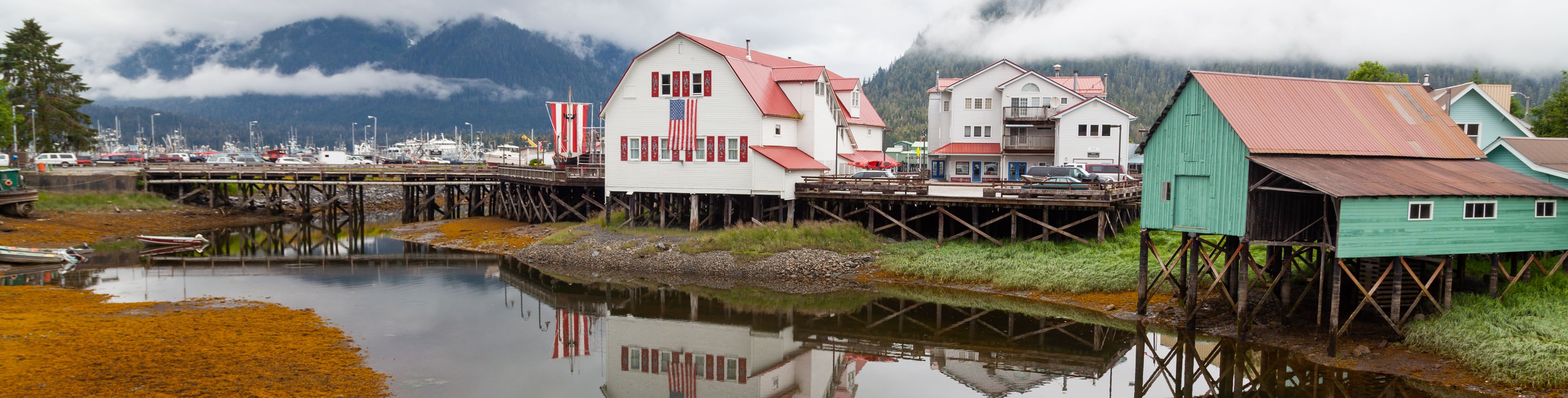 A still waterway leads between small homes with mountains in the background.