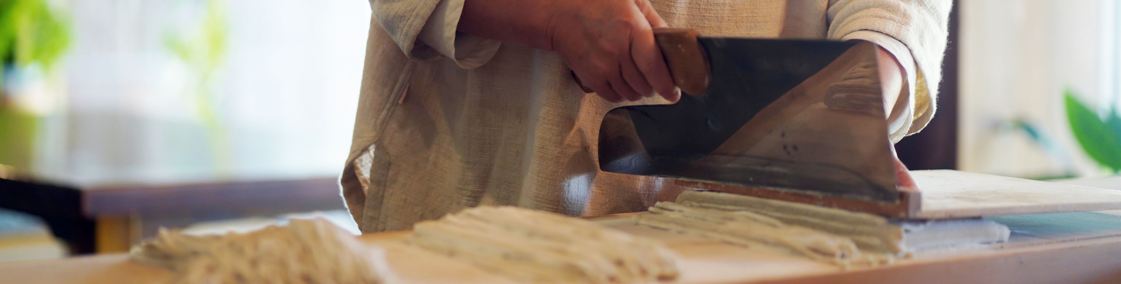 Woman using a butcher knife to thinly cut dough.