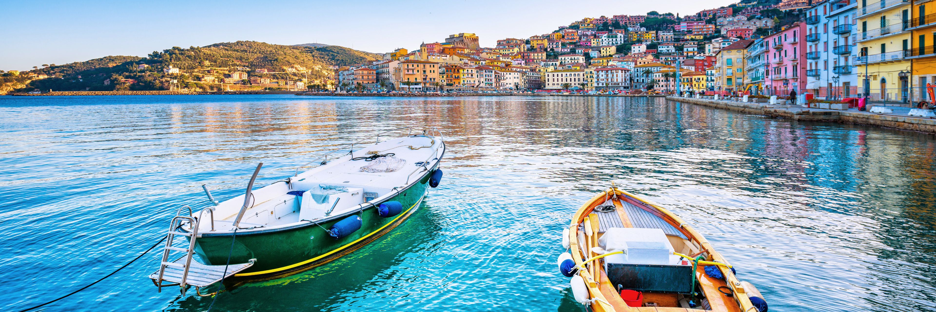 Two small boats sit on a still waterway with a row of colorful buildings in the background.