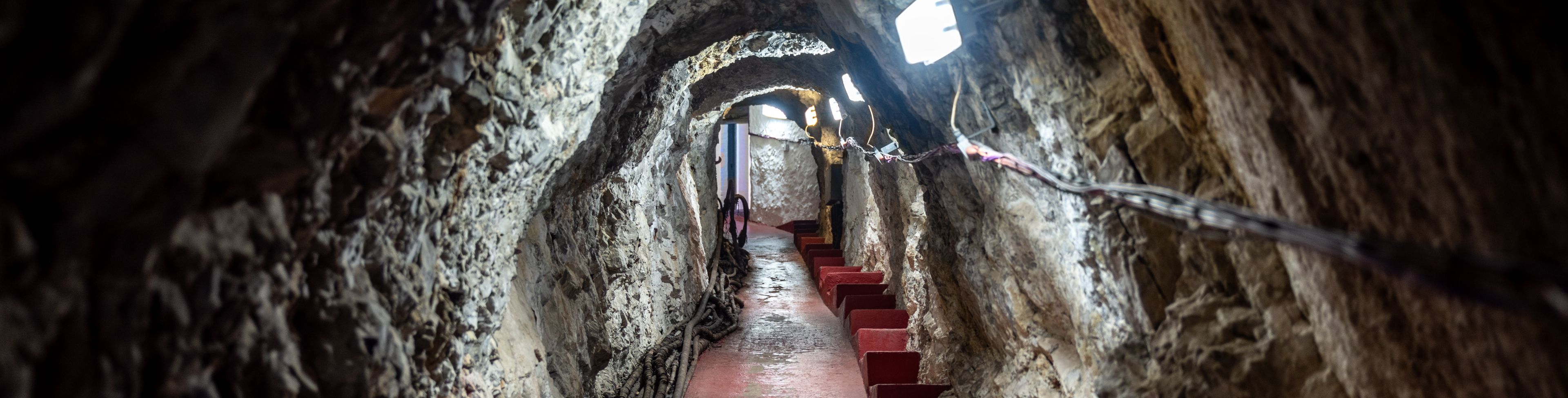 Cave tunnel with stairs leading in and out of the cave, lit with lights on the cave wall. 