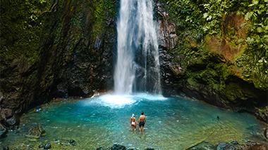 Two people standing in clear water at the bottom of a waterfall. 