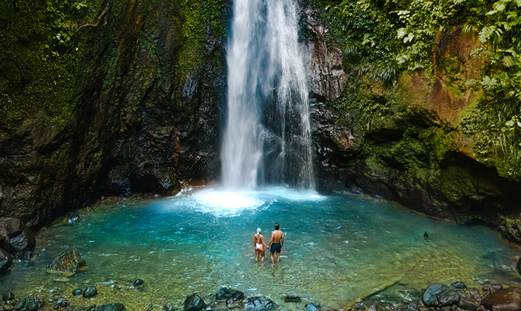 Two people standing in clear water at the bottom of a waterfall. 