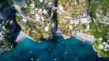 Aerial view from above, buildings and a road winding along a hillside cliff and blue ocean water at the edge of cliff. 