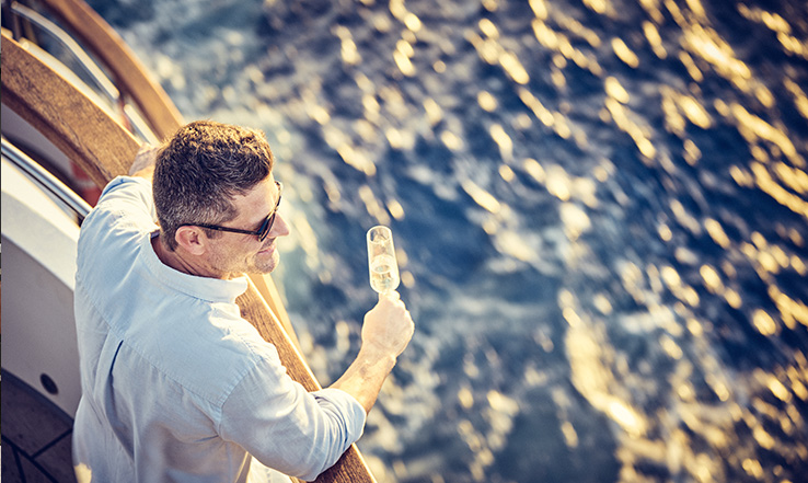 Man holding a champagne glass at the edge of a yacht over the ocean. 