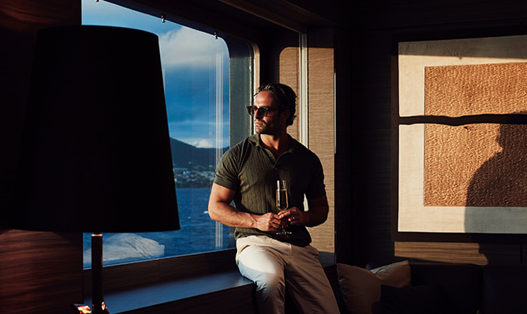 Man sitting on a table looking out the window with a drink in his hand. 