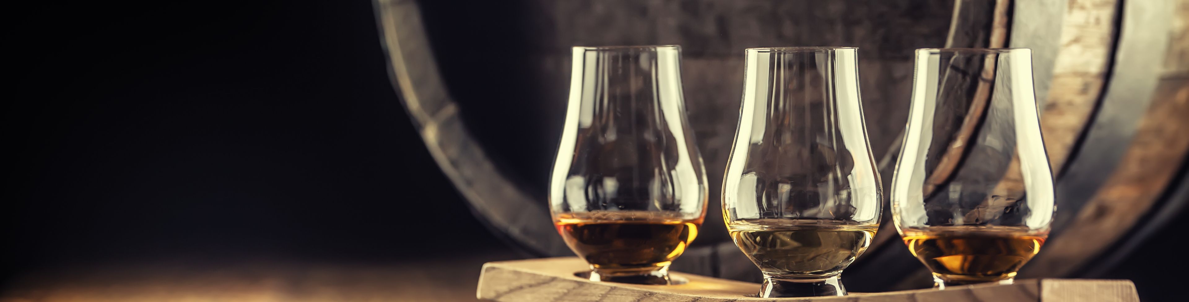 Three glasses full of different drinks to sample in front of a barrel. 