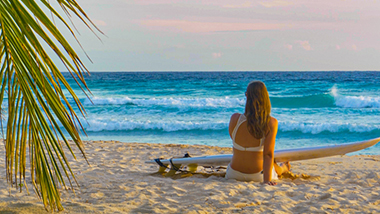 Woman on the beach with a surf board across her lap looking at the ocean. 