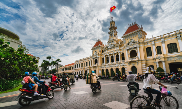Street full of people on mopeds in front of a large building. 