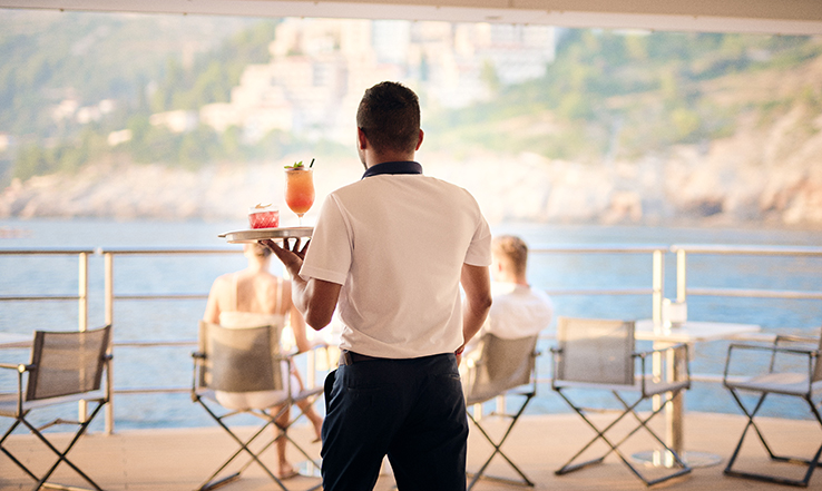 Waiter holding a tray with drinks on it with an ocean view on the back of a yacht. 