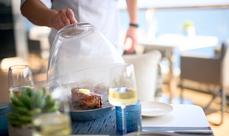 Person lifting a glass cover off of steaming food on a table with wine and plates set up. 