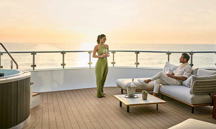 Woman standing on deck while looking at a man lounging in a chairl, the deck overlooks the ocean sunset.