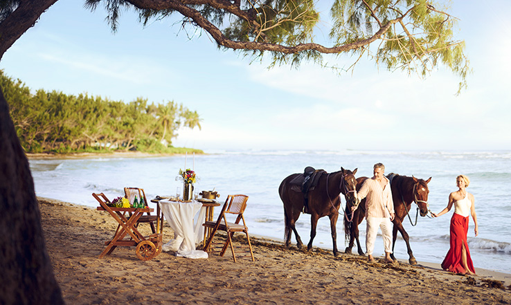 Man and woman walking horses on a beach, with a romantic table setting nearby.