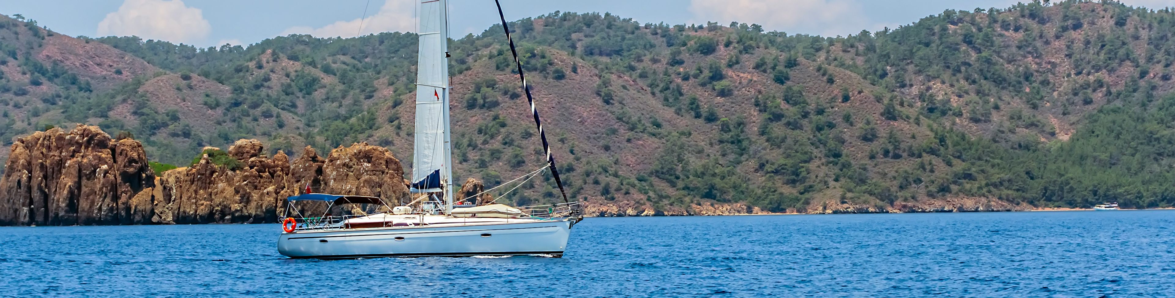 A sailboat cruises past tree lined cliffs.