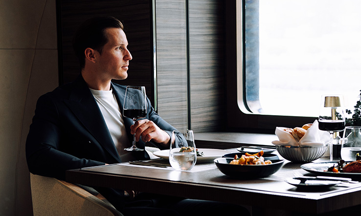 Man with wine glass in hand, staring out the window at a table.