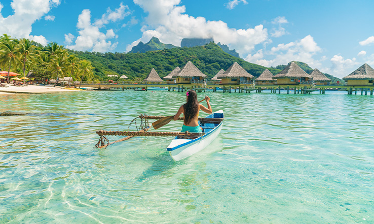 Woman rowing in a canoe toward huts above water.