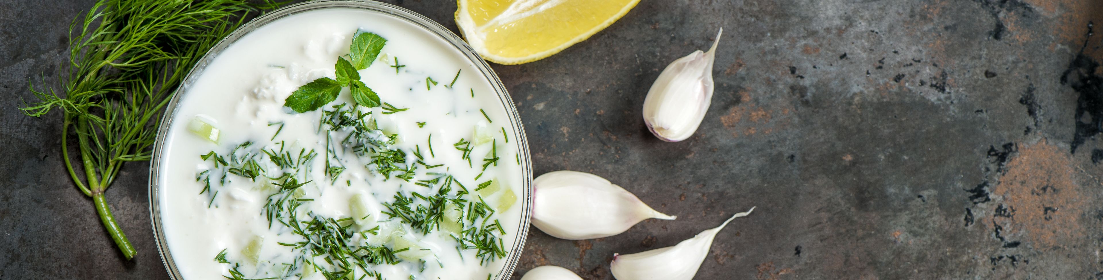 White dip in a bowl surrounded by lemons, garlic cloves, and cucumbers.