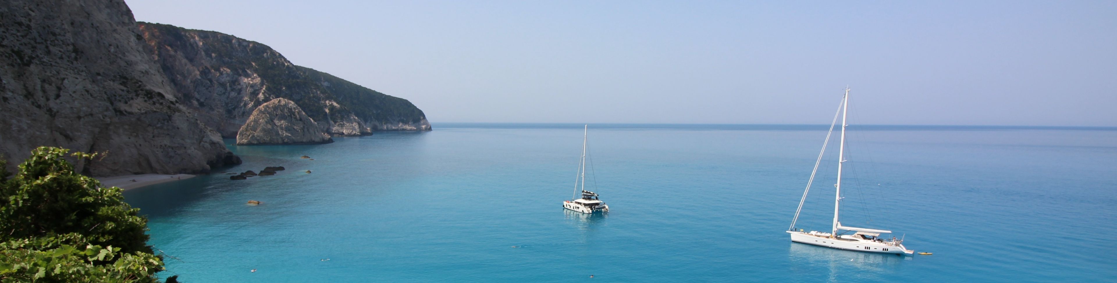 View of mountainside going into the beach and sail boats on the water in the distance.