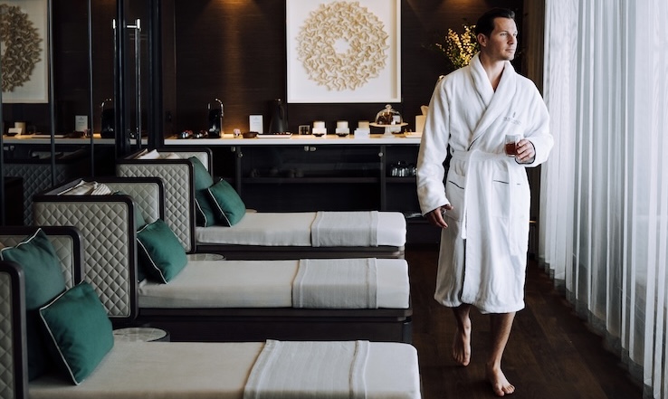 Man holding a drink, wearing a robe while walking past indoor lounge chairs.